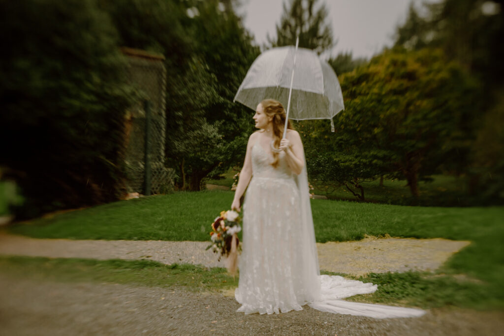 Bride in lacy dress waits under umbrella in Mitchell Grove
