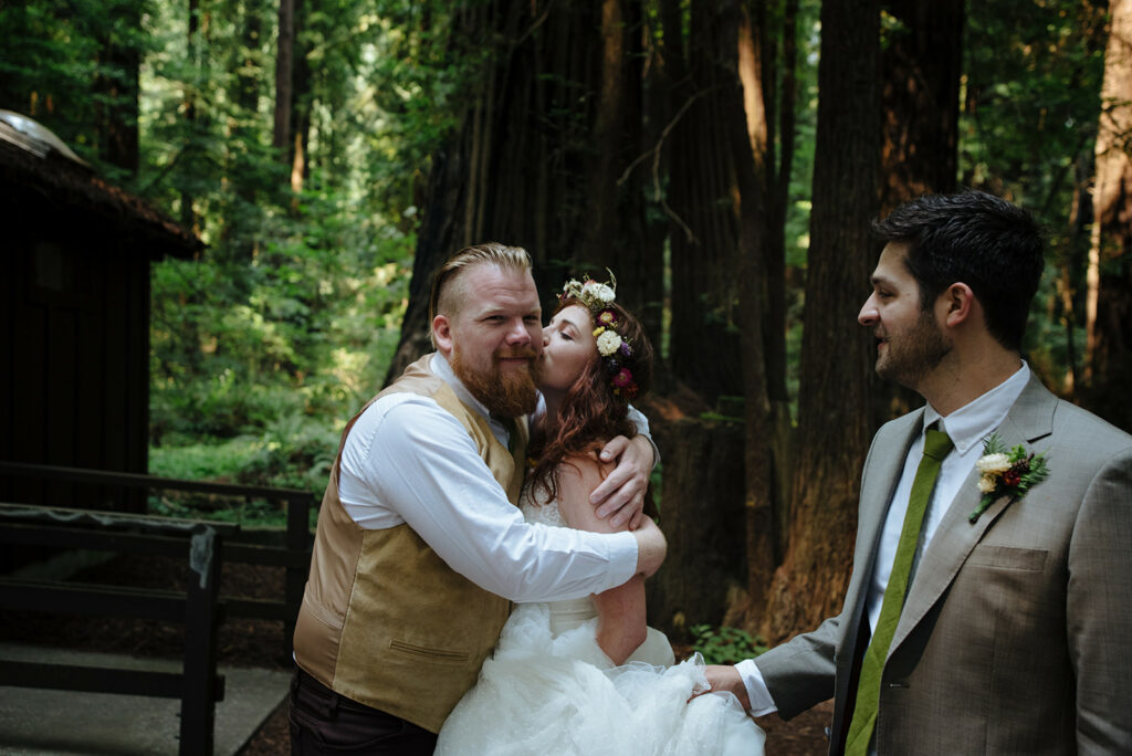 Bride in straw flower crown hugs groomsman while groom holds dress in redwoods
