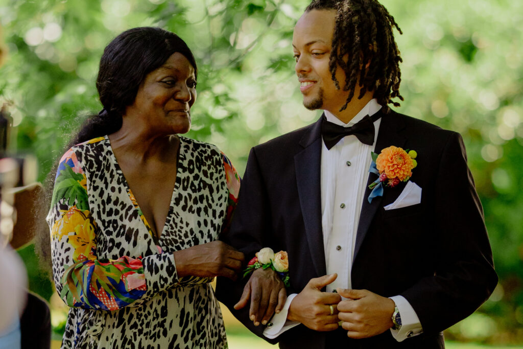 Groom with dreadlocks in black tuxedo walks down aisle with Mom in colorful floral and leopard dress