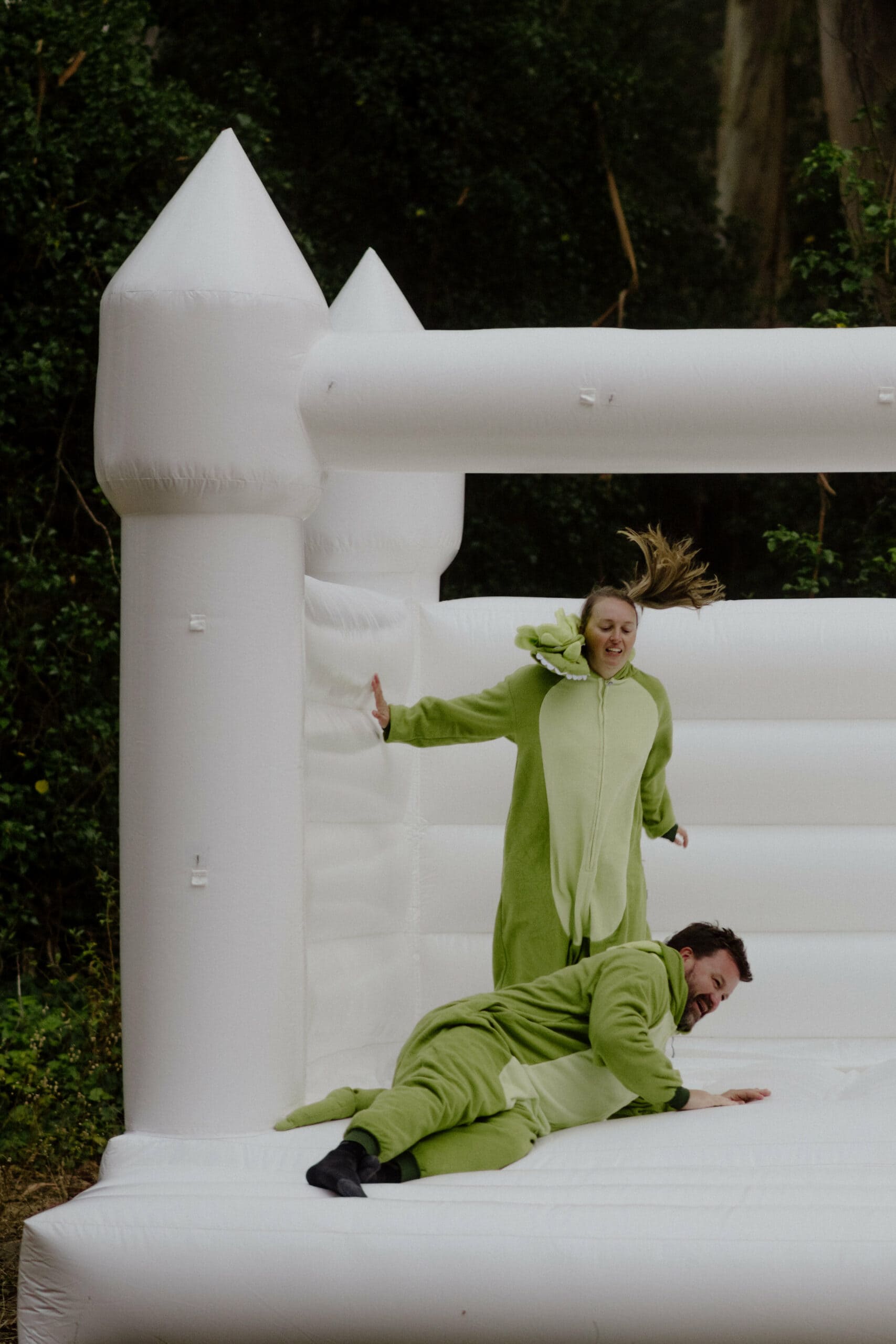 Guests in a green dinosaur onesies jump in bouncy castle at Humboldt Bay Social Club Oyster Beach wedding in Northern California