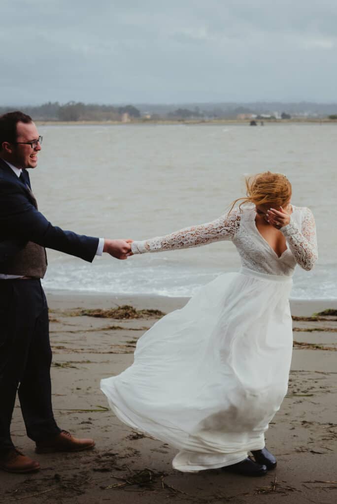 Bride hangs on to groom as her gown billows in the wind on Oyster Beach at Humboldt Bay Social Club in Samoa, California.