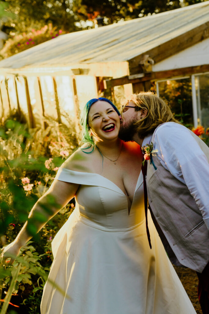 Bride with blue hair laughs while groom kisses cheek in the gardens at the Lost Whale Inn on the redwood coast in Trinidad, California.