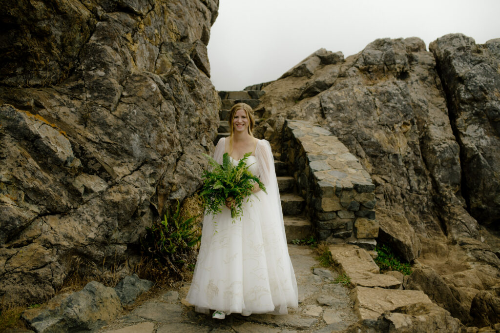 Bride with flowing cape and fern bouquet descends the stairs at Wedding Rock in Sue-meg State Park