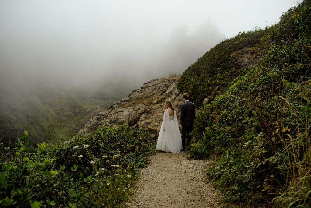 Bride with flowing sheer cape walks with groom in grey suit down trail from the top of Wedding Rock
