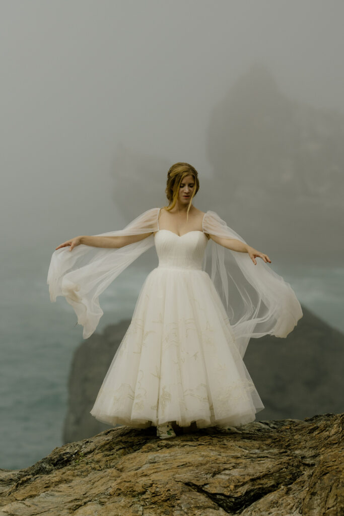 Bride lifts cape in the breeze on Wedding Rock during Northern California wedding portrait session