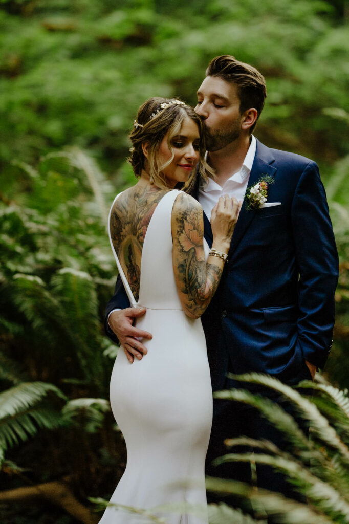 Newly married couple in navy suit and v-back fitted gown with beautiful tattoos cuddle on a fern lined trail in Redwood National Park