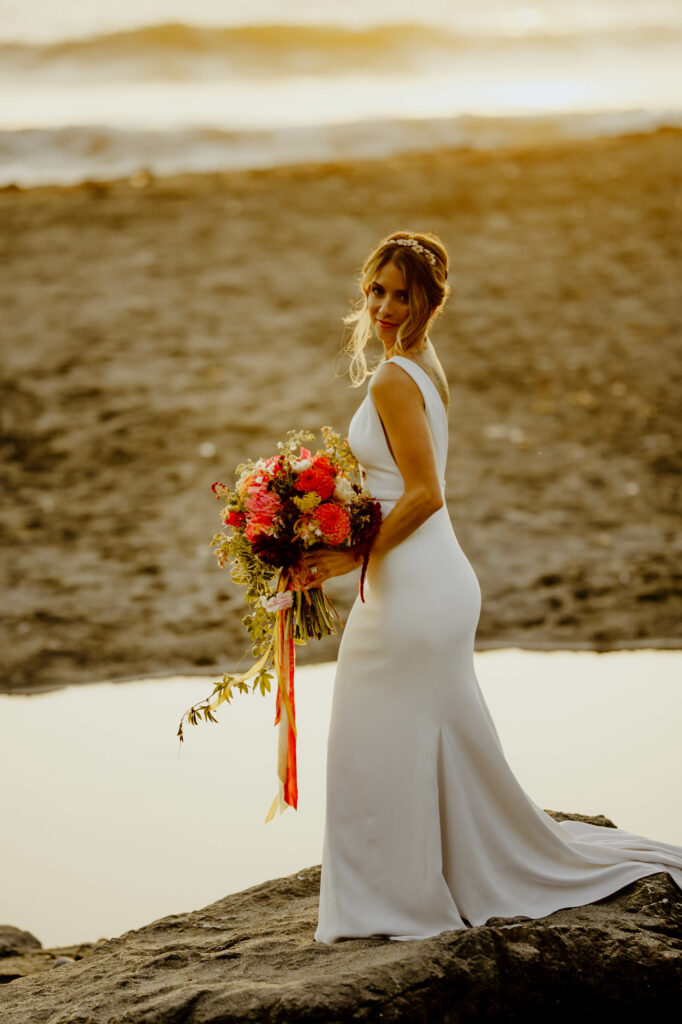 Bride holds bright red, pink, orange and cream bouquet with velvet ribbons at golden hour on Luffenholtz Beach in Trinidad, California.