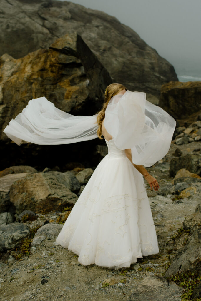 Bride stands on rocky shore in Sue-meg State Park in Trinidad, California while her cape catches the breeze