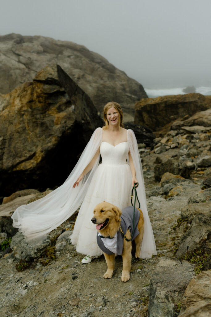 Bride in sheer floor length cape with jurassic embroidery on her gown laughs with golden retriever in grey tuxedo vest during a bridal portrait session on a rocky outcrop beside the Pacific Ocean in Trinidad, California