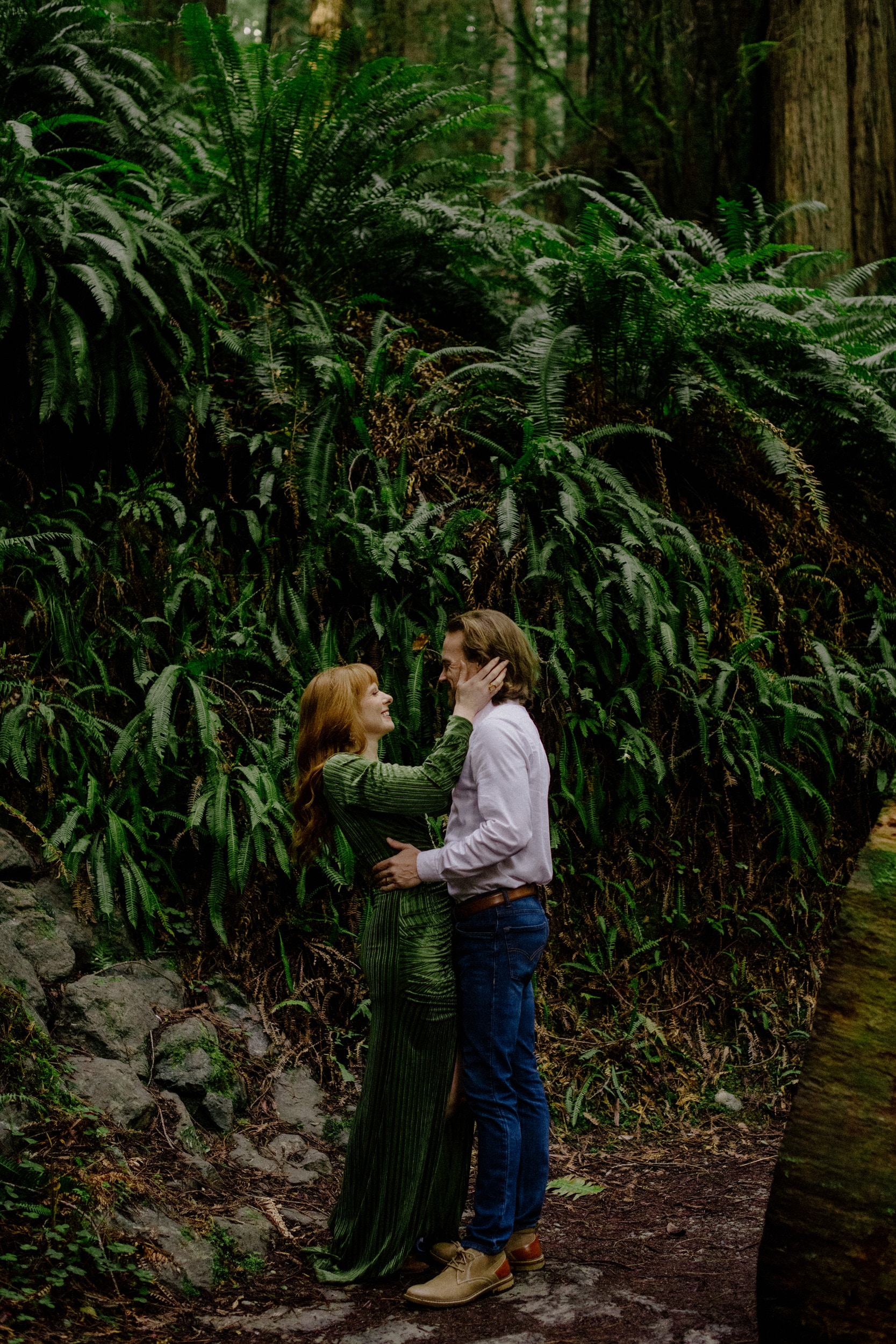 Bride in green velvet dress and vintage boots in Prairie Creek Redwoods embraces husband near a fern wall
