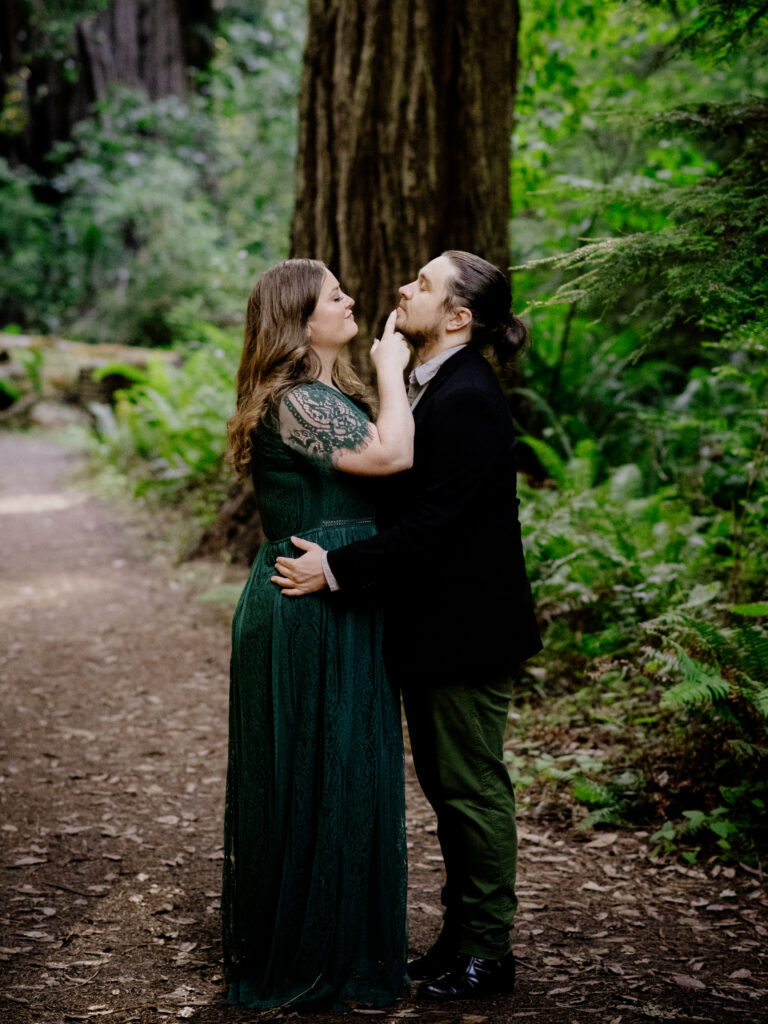 Engagement portrait of two marriers in Redwood National Park. Partner with long hair and a green lace dress places finger tip on the chin of partner in black blazer and dark green pants with hair pulled into a pony tail.