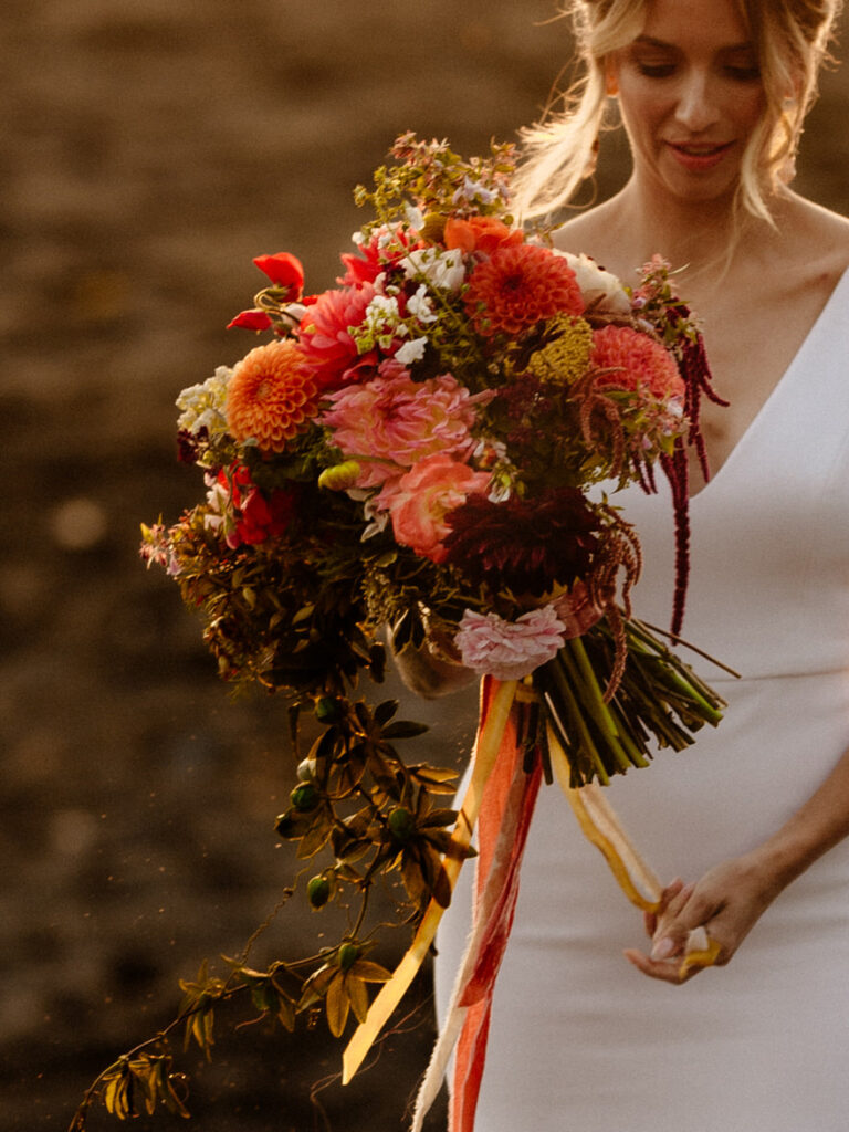 Late summer dahlia bouquet with velvet ribbon held by newly married person in fitted modern wedding gown at sunset on Luffenholtz Beach in Northern California
