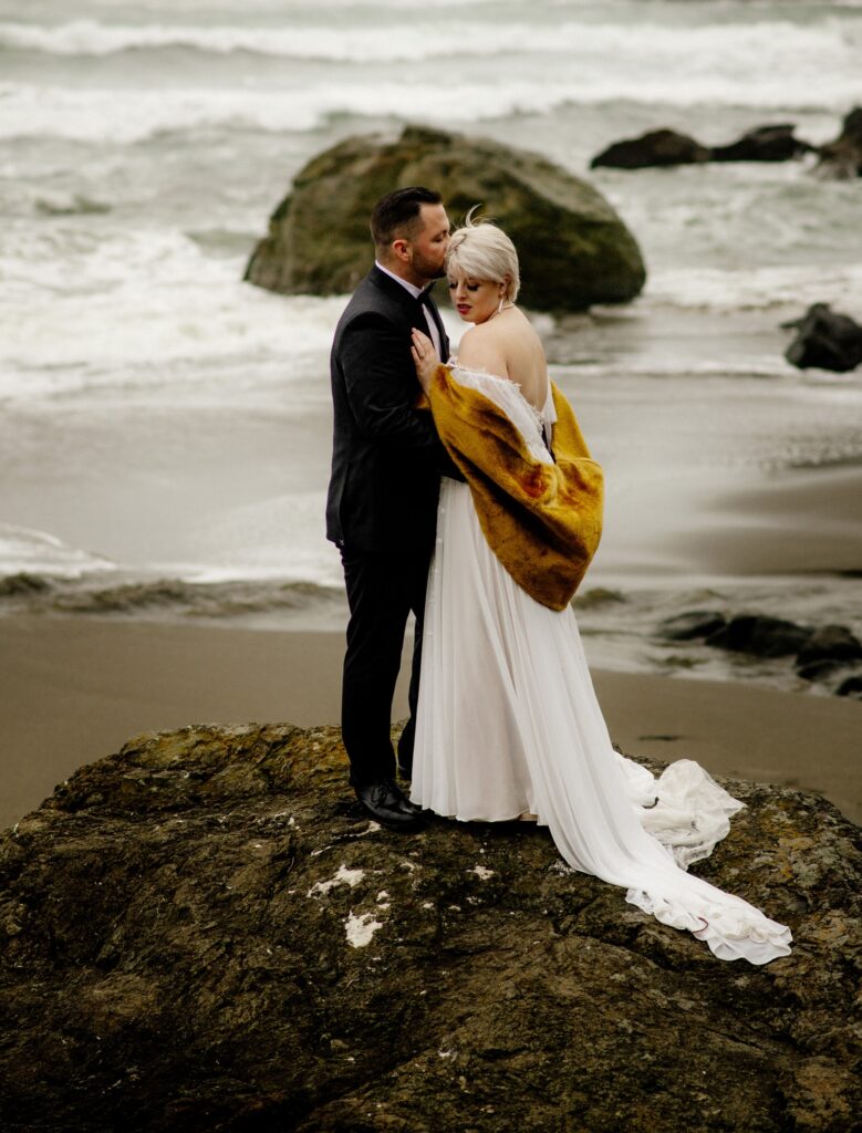 Newly married couple in formal wedding attire embrace atop a large rock on a cloudy day with Trinidad State Beach and a stormy sea in the back ground.