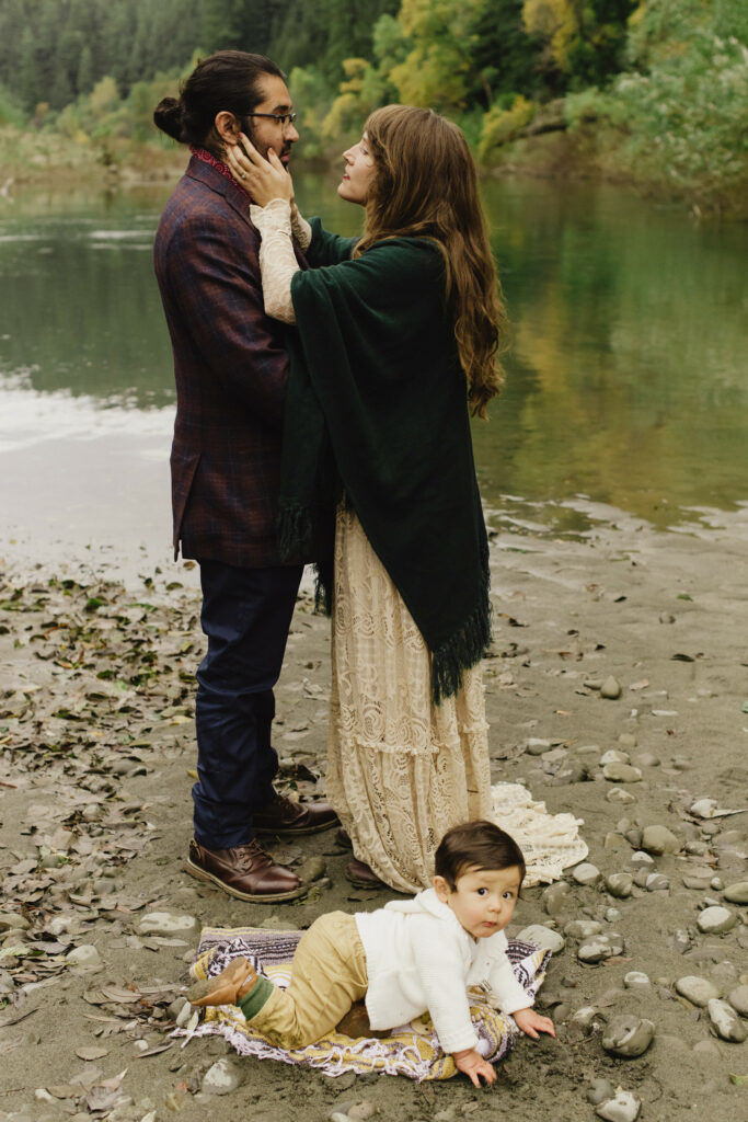 Bride in green wool cape and lace gown cradles the face of her groom who wears a purple plaid sport coat while their infant son crawls on a blanket at their feet in Williams Grove along the Avenue of the Giants in Northern California.