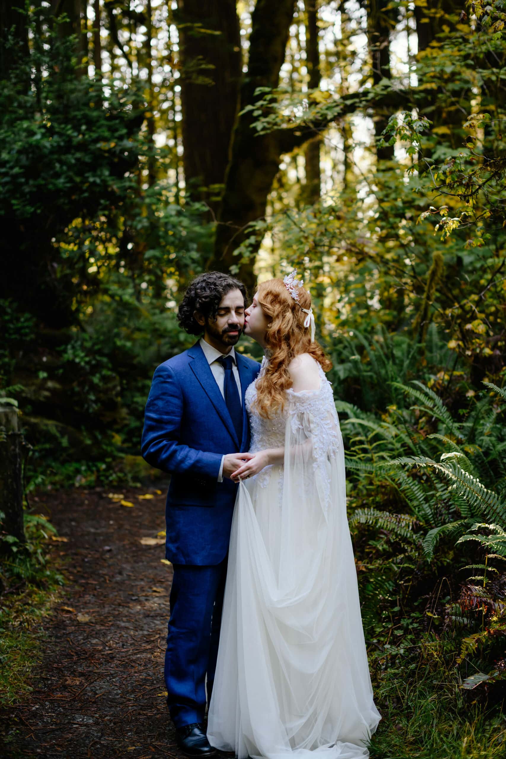Newlywed portrait in Redwood National Park in Northern California with groom in royal blue linen suit and bride wearing vintage lace gown with sheer cape and a butterfly crown.